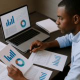 Homme analysant des graphiques et des données sur un ordinateur portable et des documents imprimés dans le cadre d’un mémoire ou d’une étude académique.